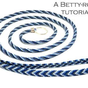 以下が含まれることがあります： 金色のリングと銀色のビーズのアクセントが付いた、青、白、グレーの編まれたランヤード。ベティ・ローズのチュートリアルです。