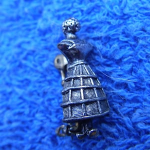 May include: A silver-toned brooch featuring a stylized figure of a woman wearing a dress with a cage-like skirt.