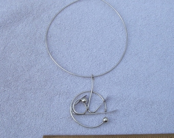 Puede incluir: Un collar de plata con un colgante circular. El colgante está hecho de alambre retorcido y tiene dos pequeñas bolas de plata.