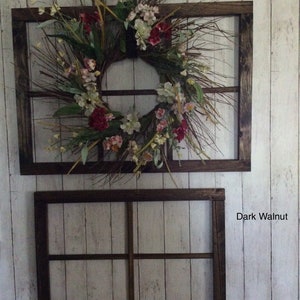 Puede incluir: Dos marcos de ventana de madera teñida de nogal oscuro están montados en una pared de madera pintada de blanco. El marco superior contiene una corona floral con acentos rojos, blancos y verdes. El marco inferior está vacío.