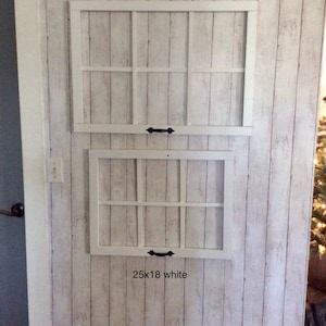 Op de afbeelding: Twee witte decoratieve raamkozijnen, één van 76x53 cm en de andere van 64x46 cm, zijn gemonteerd op een muur met een witte houtlook afwerking. Elk frame heeft een rasterpatroon en een zwarte handgreep.