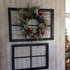 May include: Two dark-framed window decorations hang on a white, wood-panelled wall. The top frame holds a floral wreath with red, white, and green flowers. The bottom frame is empty. The wall has a rustic, farmhouse aesthetic.