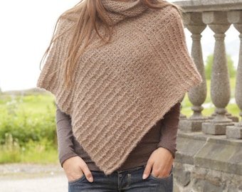 May include: A brown knitted poncho with a high neck and a diagonal stitch pattern. The poncho is worn by a person with long brown hair and is standing in front of a stone wall.