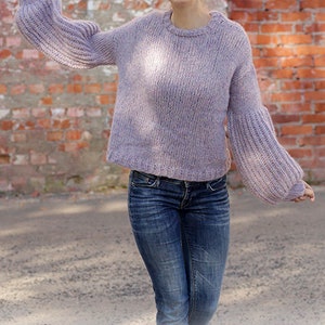 May include: A light purple knit sweater with flared sleeves and a crew neck. The model is wearing blue jeans. The background is a brick wall. The text "DROPS DESIGN" and website address are visible.