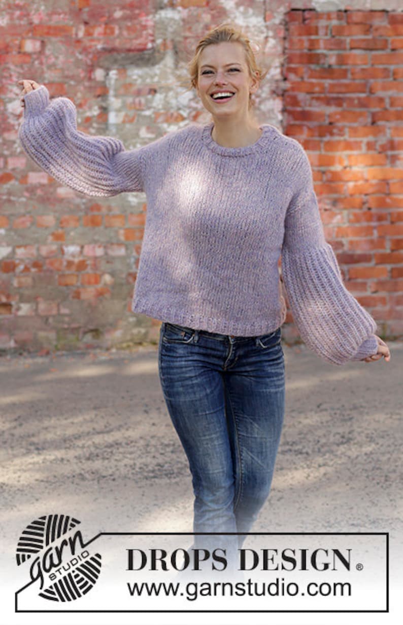May include: A light purple knit sweater with flared sleeves and a crew neck. The model is wearing blue jeans. The background is a brick wall. The text "DROPS DESIGN" and website address are visible.