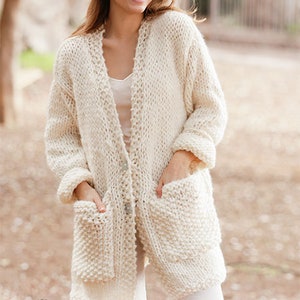 Könnte beinhalten: Ein weißer Strickcardigan mit Knopfleiste und zwei großen Taschen. Der Cardigan ist aus einem dicken Garn gestrickt und hat eine lockere Passform.