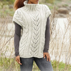 May include: A light gray, sleeveless knitted sweater with a high neck and intricate leaf-like patterns. The model is wearing a long-sleeved gray shirt underneath and blue jeans. The image includes the text "DROPS DESIGN" and "www.garnstudio.com".