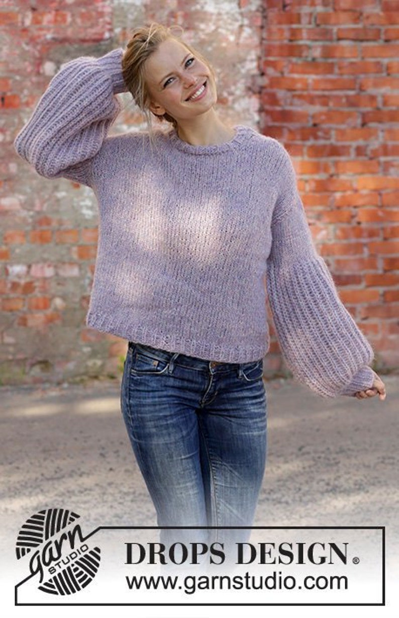 May include: A light purple knit sweater with long, voluminous sleeves. The model is wearing blue jeans. The image includes the text "DROPS DESIGN" and the website address "www.garnstudio.com".