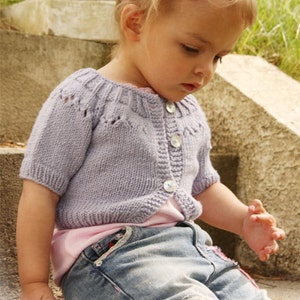 May include: A lilac knitted cardigan with short sleeves and large white buttons. The cardigan is worn over a pink top and blue jeans. A pink flower hair clip is in the child's hair. The image includes the text "DROPS DESIGN".