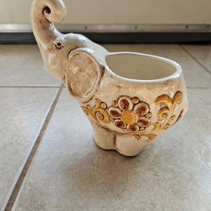 May include: Cream-colored elephant-shaped planter with a raised trunk and a decorative floral design in brown and gold. The ceramic planter has a wide opening for plants or other items.
