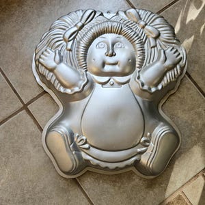 May include: A silver-colored Cabbage Patch Kids cake pan. The pan is shaped like the doll, with molded details of the face, hair, bows, arms, and legs. The pan has a loop for hanging.