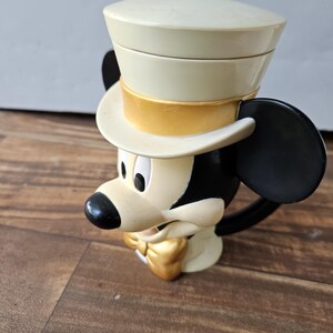 May include: A vintage Mickey Mouse mug featuring a cream-colored top hat with a gold band, black ears, and a gold bow tie. The mug is primarily cream and black, with a black handle.