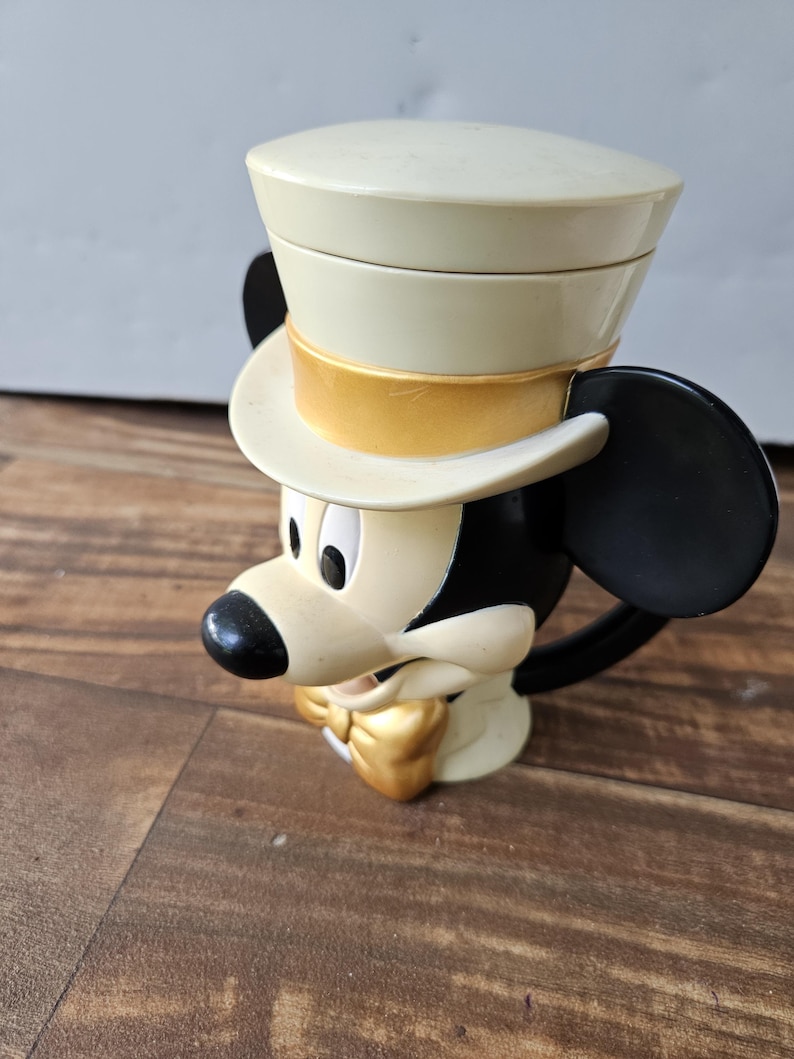 May include: A vintage Mickey Mouse mug featuring a cream-colored top hat with a gold band, black ears, and a gold bow tie. The mug is primarily cream and black, with a black handle.