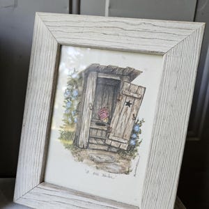 May include: A framed illustration of an outhouse with a wooden door featuring a black star cutout. The drawing includes blue flowers, a small red book inside, and the handwritten text "a one holer" at the bottom, set in a light wood frame.