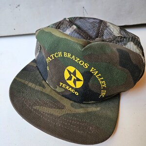 May include: Camouflage trucker hat with a yellow star and the text "Patch Brazos Valley, Inc. Texaco" on the front.