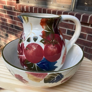 May include: A white ceramic pitcher and bowl set with a fall leaf and apple design. The pitcher has a handle and the bowl is round. Both pieces have a green, red, blue, and yellow design.
