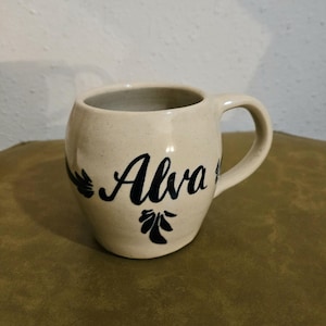 May include: Cream-colored ceramic mug with a rounded body and a handle. The word "Alva" is written in elegant black script on the front, accompanied by decorative floral accents. The mug has a rustic, handmade appearance.
