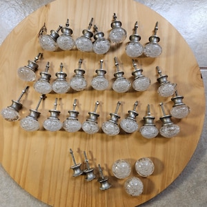 May include: A collection of 25 clear glass cabinet knobs with a silver finish. The knobs have a textured, frosted glass surface and are arranged on a wooden surface.