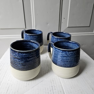 May include: A set of four ceramic mugs featuring a two-tone design with a deep blue, textured upper section and a smooth, off-white base. Each mug has a cylindrical shape with a handle on the side and visible horizontal ridges along the exterior.