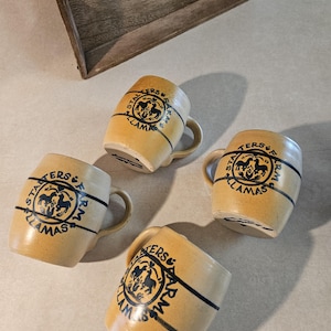 May include: Four tan ceramic mugs with black and blue accents. Each mug has a handle and a circular emblem with the words "Starters Farm Llamas" and a llama graphic. The mugs are arranged on a light-colored surface.