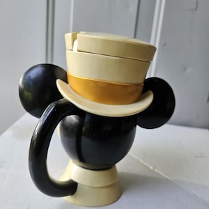 May include: A black and cream-colored Mickey Mouse-shaped mug with a handle. The mug features a cream-colored top hat with a gold band. The ears are black, and the base is cream-colored.