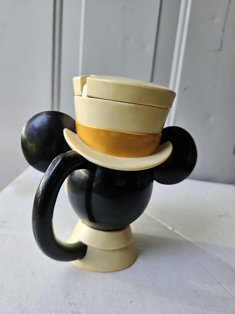 May include: A black and cream-colored Mickey Mouse-shaped mug with a handle. The mug features a cream-colored top hat with a gold band. The ears are black, and the base is cream-colored.