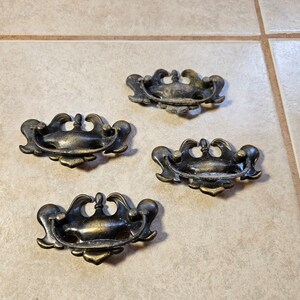 May include: Four antique brass drawer pulls with ornate, curved designs. The pulls have a dark, aged patina, suggesting vintage or reclaimed hardware. Each pull features a central handle and decorative flourishes.