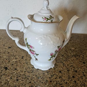 May include: White porcelain teapot with a floral design featuring pink roses and green leaves. The teapot has a gold trim around the lid and base.