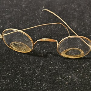 May include: A pair of round, gold-rimmed glasses with clear lenses. The glasses are slightly dirty and have a vintage look.