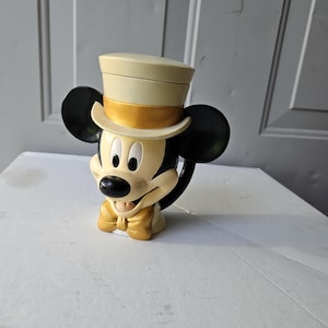 May include: A Mickey Mouse mug with a cream-colored top hat and a gold bow tie. The mug features Mickey's iconic black ears and a black handle. The mug is sitting on a white surface.