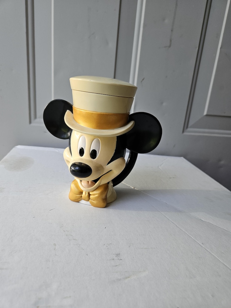 May include: A Mickey Mouse mug with a cream-colored top hat and a gold bow tie. The mug features Mickey's iconic black ears and a black handle. The mug is sitting on a white surface.