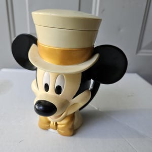 May include: A Mickey Mouse-shaped mug with a cream-colored top hat and a gold bow tie. The mug has black ears and a black nose. The mug is made of ceramic and is a collectible item.