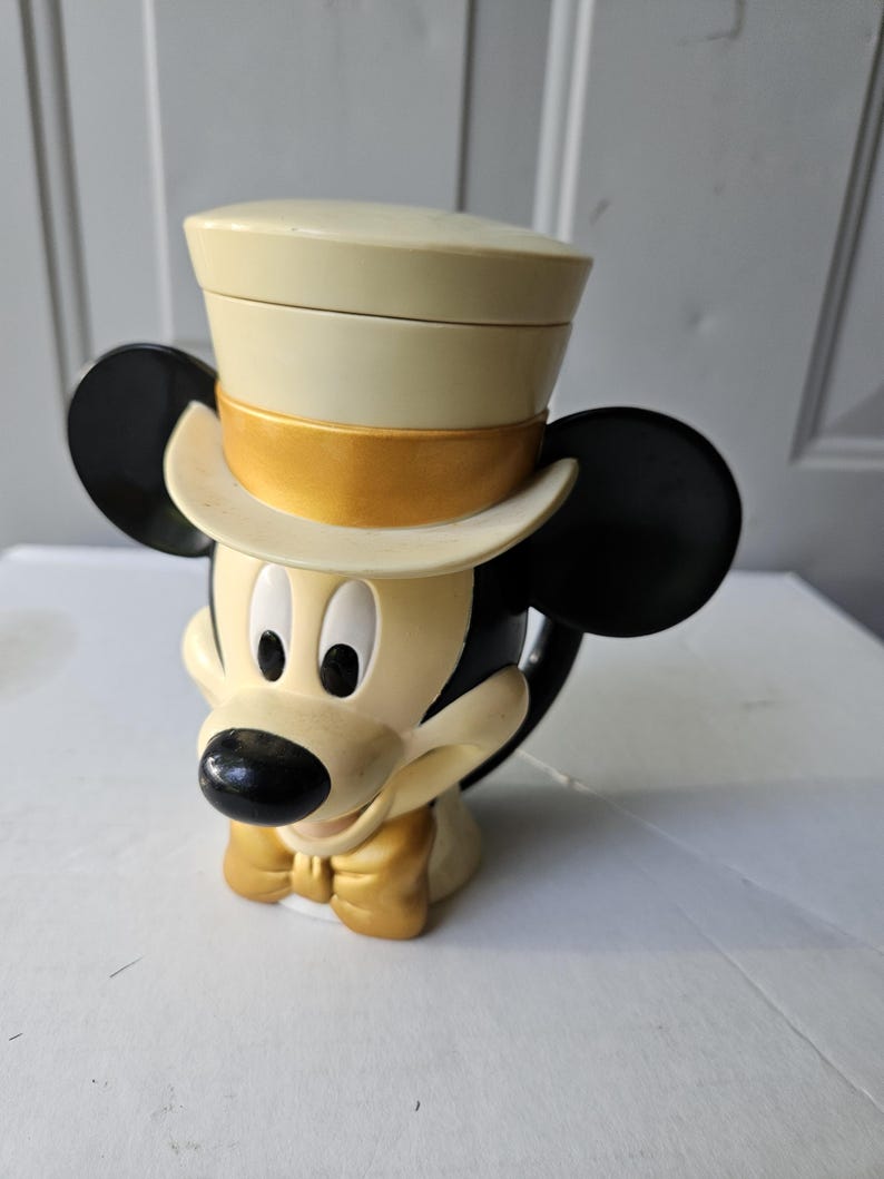May include: A Mickey Mouse-shaped mug with a cream-colored top hat and a gold bow tie. The mug has black ears and a black nose. The mug is made of ceramic and is a collectible item.