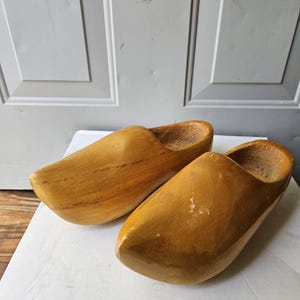 May include: A pair of wooden clogs, showcasing a light brown hue with visible wood grain. The clogs have a rounded toe and a hollowed-out interior, suggesting traditional footwear. The clogs are resting on a white surface.