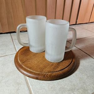 May include: Two frosted glass beer mugs with handles on a wooden coaster.