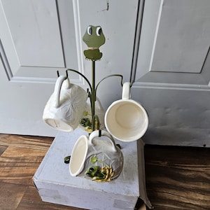 May include: A whimsical mug tree with a frog-themed design. The metal tree holds four white ceramic mugs, each adorned with a frog and lily pad motif. The tree is topped with a frog cutout. The mugs are displayed on a cardboard box.
