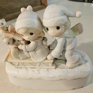 May include: A ceramic figurine depicting two children in winter attire on a sled. The children are wearing white hats and coats, with one pushing the sled. The sled is decorated with small white cat figurines. The base is sculpted to resemble snow.