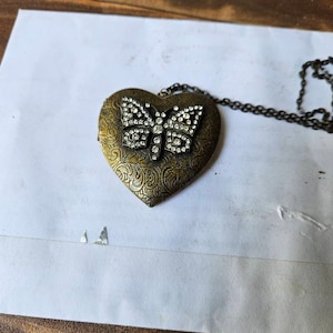 May include: A gold heart-shaped locket with a butterfly design. The butterfly is adorned with clear rhinestones. The locket is attached to a black chain.