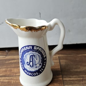 May include: White ceramic pitcher with a gold rim and a blue and white logo that reads "Aquarena Springs San Marcos, Texas" with a seahorse inside a seashell.