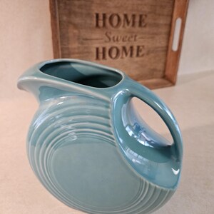 May include: A turquoise ceramic pitcher with a large handle and a rounded body. The pitcher has a series of concentric ridges around its base. Behind the pitcher is a wooden tray with the words "HOME Sweet HOME" engraved on it.