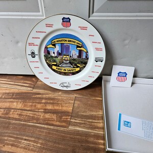May include: A white ceramic plate with a gold rim features a colorful illustration of a train and the text "The Houston Service Unit 2003 First in Safety 2003". The plate is surrounded by a circle of red text listing various locations, including "Settegast Terminal", "Englewood Terminal", "Galveston", "Sugar Land", "Baytown", and "Eagle Lake".