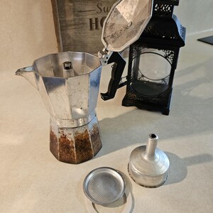 May include: An aluminum stovetop espresso maker with its lid open, showing the interior. The base has rust-colored staining. A filter, gasket, and funnel are next to the coffee maker. A black metal lantern and a wooden sign are in the background.