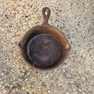 May include: A small, cast iron skillet with a loop handle. The skillet is rusty and has a dark brown color.