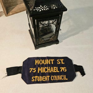 May include: A black metal lantern with glass panels and decorative cutouts, next to a navy blue banner with gold text that reads "MOUNT ST. 75 MICHAEL 76 STUDENT COUNCIL."