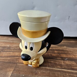 May include: A Mickey Mouse figurine wearing a cream-colored top hat with a gold band. The figurine has black ears, a black nose, and a gold bow tie. The figure is cream-colored with black accents.