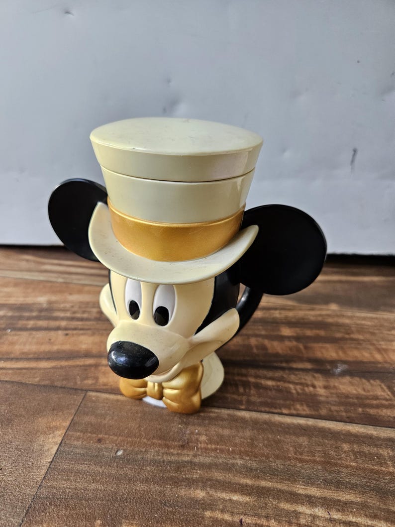 May include: A Mickey Mouse figurine wearing a cream-colored top hat with a gold band. The figurine has black ears, a black nose, and a gold bow tie. The figure is cream-colored with black accents.