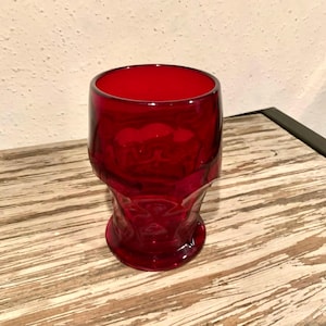 May include: A ruby red glass with a skull design. The glass has a wide opening and a thick base. The glass is made of a transparent material, allowing light to pass through. The glass is sitting on a wooden surface.