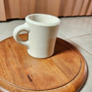 May include: A white ceramic mug with a handle, sitting on a round wooden coaster.