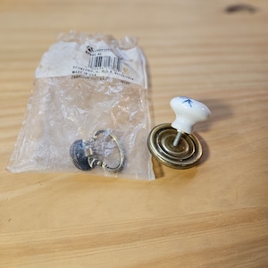 May include: Two cabinet knobs, one with a white ceramic base and blue floral design, and the other with a brass base and a decorative ring. Both knobs are in a clear plastic bag with a label that reads "Made in U.S.A."