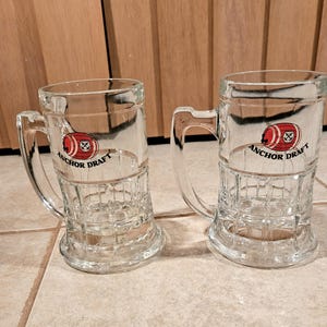 May include: Two clear glass beer mugs with handles. Each mug has a red and white football logo with the words "Anchor Draft" printed below. The mugs are sitting on a tiled surface.